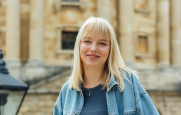 Eleanor Miller wearing a blue jacket stands in front of the Radcliffe Camera on a sunny day
