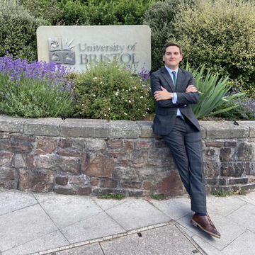 An image of Jose Rojas Alvarado, wearing a suit and standing outside next to a University of Bristol sign. 