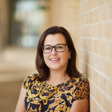 Laura smiling outside, leaning against a brick wall. She has shoulder-length brown hair, is wearing brown glasses and a gold and black patterned top. 