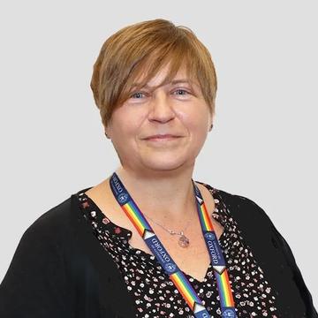 An image of Louise smiling against a plain backdrop. She has short blonde hair, and is wearing a black cardigan and patterned shirt with a rainbow lanyard. 