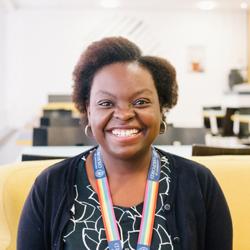 Machilu smiling at the camera, sitting on a yellow seat. She is wearing a rainbow lanyard and a patterned black and white top. 