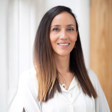 Nina smiling at the camera, dressed in a white top. She has long brown hair and is standing against a white background with a wooden door. 