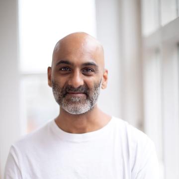 Sanjiv is smiling at the camera with his arms folded. He is wearing a white T-shirt and standing against a white backdrop with windows. 