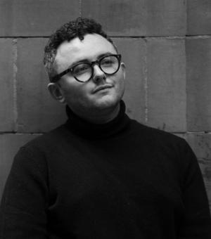 A black and white photograph of Jacob, wearing a black turtleneck and black rimmed glasses.